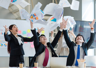 Wall Mural - Millennial Asian young professional successful male female businessmen businesswomen in formal suit laughing smiling throwing paperwork documents up in air celebrating together in company office