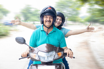 Happy senior indian couple wearing helmet riding motor scooter on road. Retirement life, Adventure and travel, Closeup, front view.