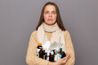 © sementsova321 - Portrait of ill beautiful caucasian woman wearing jumper and scarf holding pills, standing isolated over grey background, looking at camera with pout lips and crying.