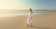 Woman Walking At Sandy Beach On Summer Vacation In Fuerteventura Canary Islands Spain. Adult Girl In White Dress Sunglasses Enjoying Holiday.
