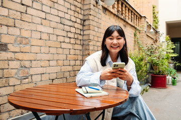 Wall Mural - Young asian girl using smartphone and smiling at camera while sitting in cafe