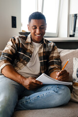 Canvas Print - Young african man writing down notes while sitting on sofa at home