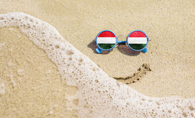 Wall Mural - Sunglasses with flag of Hungary on a sandy beach. Nearby is a sea lightning and a painted smile. Travel and vacation concept for Hungarians