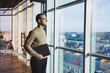 © DSMT - A cute young man in a brown sweater and glasses is standing near a large window in the office with a laptop in his hands while working in the office. A young freelancer works remotely.