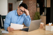 © Prostock-studio - Confused asian mature man sitting in front of laptop, reading papers and touching his head, copy space
