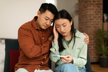 Upset mature asian husband comforting his depressed young wife with negative pregnancy test, sitting on sofa at home