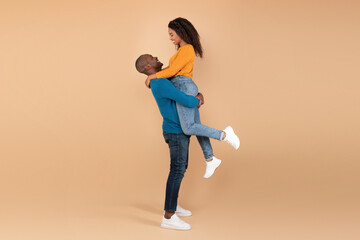 Loving mature black man lifting his wife and holding her on peach studio background, profile side view, free space