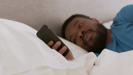 Wall Mural - Gadgets in modern life. Young african american man web surfing on smartphone, lying in bed, tracking shot, free space