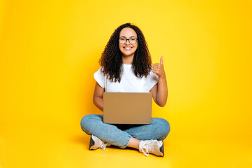 Full-length photo of positive pretty, proud latino or brazilian woman, in casual wear, sits with an open laptop in lotus position on isolated yellow background, smiles at camera,shows thumb-up gesture