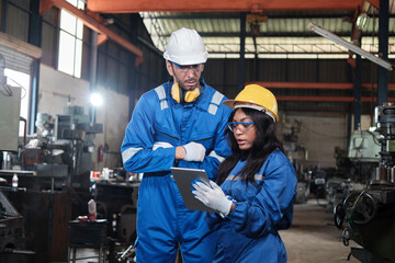 Wall Mural - Two industrial workers in safety uniforms and hardhats, male manager, and Black colleague work with tablet to check metalwork machines in manufacturing factory. Professional production engineer team.