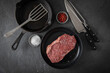 Entrecote Steak roh vom Rind mit Meersalz und roter Pfeffer auf Teller bei Vorbereitung in Küche mit Messer, Fleischgabel, Gusseisern Pfanne und Pfannenwender auf Schiefer Stein Hintergrund Schwarz