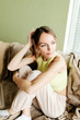 © Mila - A young beautiful girl with blond hair gathered in a ponytail sits on a sofa by the window. Beautiful young woman in a green top relaxing on the sofa