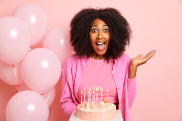 Portrait shot of positive emotional woman with surprised expression, holds delicious cake with burning candles, prepares for the party, poses over pink wall