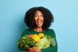 Woman holds a lot of vegetables, she likes healthy lifestyle and nutrition, so she smiles happily and ready to act