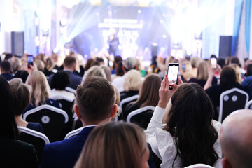 Guests in evening attire sit in a spotlighted lobby and look at the stage. Girl filming an event on the phone