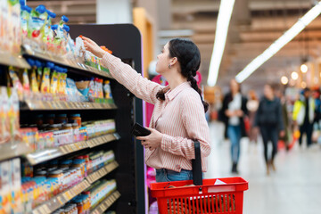 Wall Mural - Side view of young Caucasian woman reaches hand to top shelf, holds cellphone and cart. Shelves with food in background. Concept of shopping in grocery store