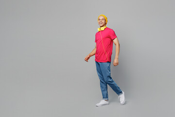 Wall Mural - Full body side view smiling happy fun cheerful young man of African American ethnicity 20s wear pink t-shirt yellow hat headphones walking going look camera isolated on plain grey background studio