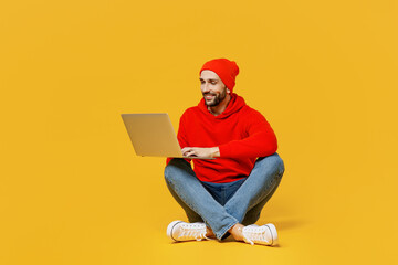 Full body young smiling programmer happy caucasian man in red hoody hat sitting hold use work on laptop pc computer isolated on plain yellow color background studio portrait People lifestyle concept