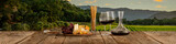 Fototapeta Kawa jest smaczna - Decanter and two glasses with delicious red wine, grape and cheese appetizers standing on wooden table over beautiful nature landscape of fields and forest.
