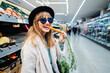 Stylish fashion smiling woman eating fresh carrot in the supermarket store during selecting fresh products. Veganuary month, Healthy eating diet, go vegan. Selective focus. Copy space.