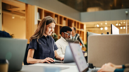 Portrait of Caucasian Inspired Young Woman Smiling and Using Laptop in Bright Contemporary Office. Client Advisor Answering Customer Support emails. Team Working on Problem Solving in the Background