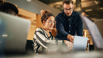 Two Colleagues Working On Laptop in Office. Female Hispanic Help Desk Coordinator Collaborates with White Male Customer Service Agent, They Discuss a Project, Chat, Smile. Teamwork Collaboration