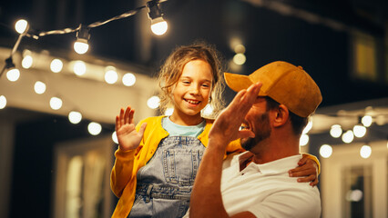 Wall Mural - Cinematic Shot of a Father Holding His Small Daughter in His Arms, Helping Her to Fix a Lightbulb in a Backyard Lights Installation. Father and Daughter High Five and Celebrate Successful Repair.