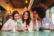 © Jose Calsina - Three ladies smiling and having fun using a cellphone to send text messages at restaurant or coffee shop, A group of happy young women laughing using their mobile phone app to share photos. High
