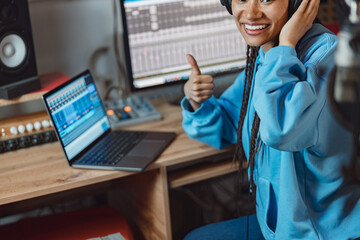 Wall Mural - Close-up of pretty African anchorwoman with headphones showing thumb up while broadcasting on radio in recording studio