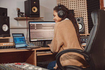 Wall Mural - African cute woman blogger records online podcast using laptop, headphones and microphone in professional music studio