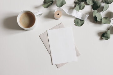 Blank greeting card, invitation mockup scene with dry eucalyptus tree branches and ribbon. Cup of coffe, white table background. Feminine breakfast composition. Flat lay, top view. Elegant stationery.