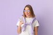 © Pixel-Shot - Young woman in eyeglasses using mobile phone on lilac background