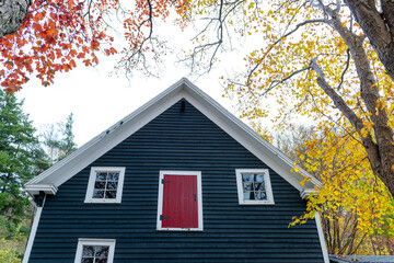 A navy blue country style farm house with a single red wooden door, two glass multi-pane windows on the exterior of the building. The trim on the shed is white colored. The shed is surrounded by trees