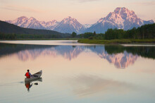 A Man Canoeing On A Calm River At Sunrise With Huge Snow Covered Mountains In The Background.