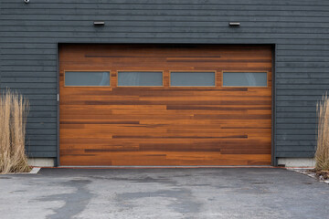 A modern brown faux wooden exterior garage door with four small horizontal glass windows. The modern door is on a luxury dark grey contemporary house with a concrete driveway. 