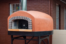 An Exterior Of A Gas Clay Pizza Oven On Black Metal Legs On A Restaurant Patio. There Are Bricks Surrounding The Opening Of The Italian Equipment.  A Temperature Gauge Is In The Middle Of The Oven.