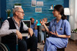 © DC Studio - Female health specialist explaining rehabilitation therapy to an elderly patient in a wheelchair in the waiting room of a nursing home. Man with disability listening to therapist, specialized care.