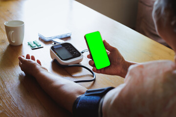 Senior asian woman is measuring blood pressure and heart rate with digital pressure gauge electric tonometer by herself at home and record result in the smartphone with chroma key screen.