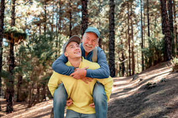 Wall Mural - Handsome boy carries his grandfather on shoulders as he walks happily in the forest. Smiling multigeneration family couple enjoying mountain and nature together. The new generation helps the old one