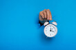 © yavdat - female hand holds pink alarm clock through a paper hole in blue background.