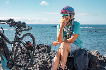 Canvas Print - Happy senior woman sitting at the beach resting close to her electric bicycle eating an apple. Authentic retirement living and healthy lifestyle concept. Horizon over the sea