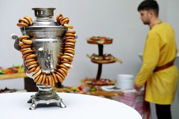 Wall Mural - Banquet in Russian style. On the table is a Russian samovar with bagels as decoration. A young man in a Russian national costume stands in the background