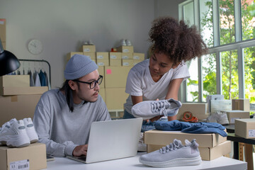 Mixed race couple of Asian man and African-American woman online seller confirming orders from customer on laptop computer. E-commerce business owner working together in store warehouse.