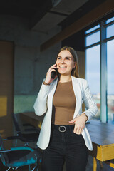 A smiling young woman in a jacket stands in the office and happily talks on the phone. A young manager works in the office and manages the business remotely