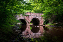 A Historic Double Arch Stone Bridge Reflects Onto The Still Stream Waters Below Surrounded By Lush Dense Green Forests During A Cloudy Summer Day. 
