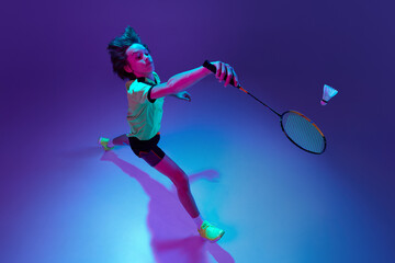 Wall Mural - Top view. Portrait of teen boy in uniform playing badminton over blue purple background in neon ligth