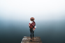 Young Woman Wearing Hat And Mountain Clothes Standing Alone On Pier And Looking At View. Mist Over Water By The Lake. Foggy Air. Empty Place For Sentimental, Inspirational Text, Quote Or Sayings.
