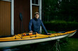 nordic finnish caucasian white outdoor man taking care of his kayak that is yellow wearing blue in front of an old vintage barn during summer evening wearing a cap in finland
