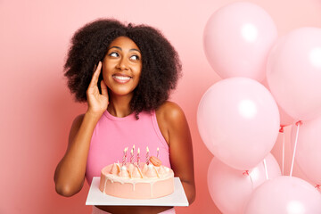 Beautiful pensive young woman with bushy hair poses with birthday cake near balloons, enjoys celebration, isolated over pink wall