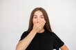 Portrait of shocked young woman covering mouth with hand over white background. Caucasian lady wearing black T-shirt looking at camera in fear. Shock and fear concept